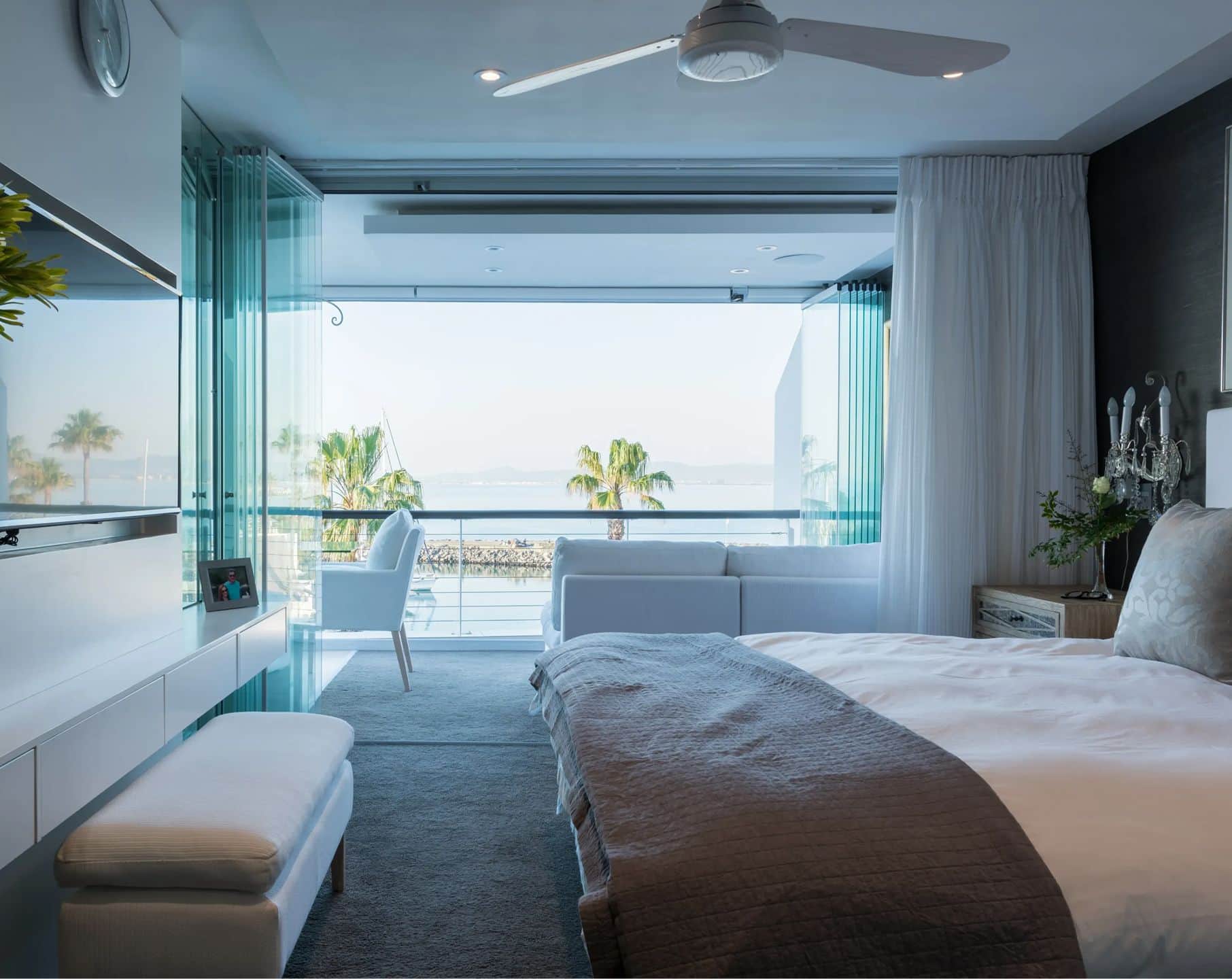 Main bedroom with folding glass doors open to a balcony overlooking palm trees and the water