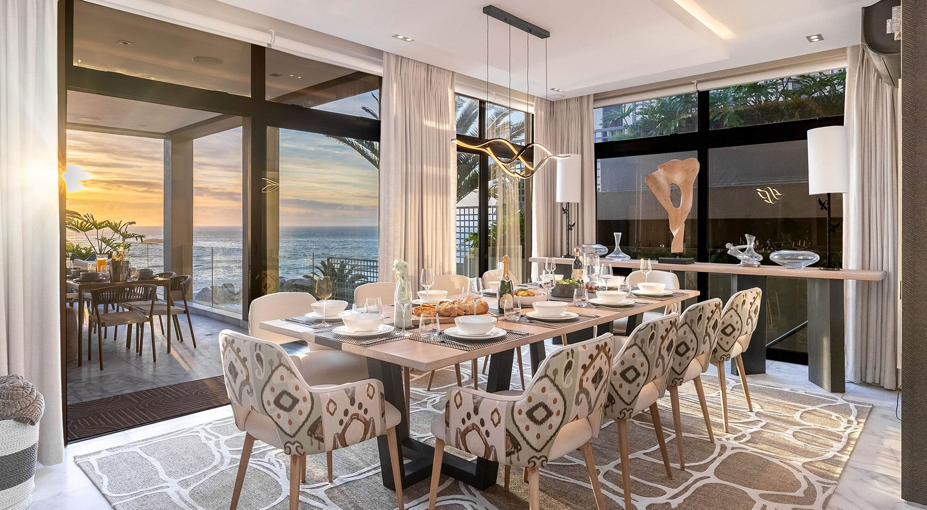 Dining room at sunset with patterned chairs around a long table and glass doors open to an ocean view