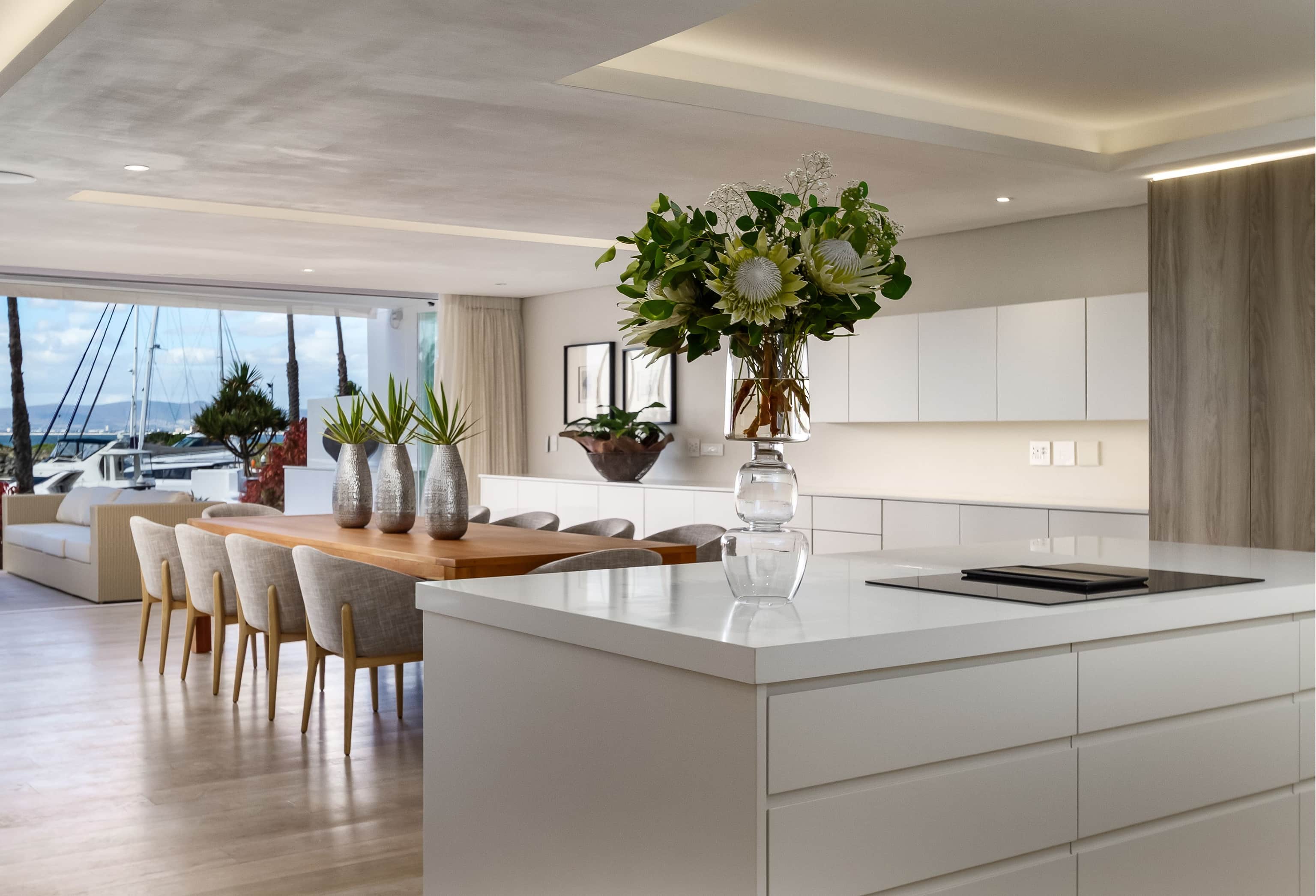 White kitchen island with a protea arrangement beside a dining table looking toward the marina