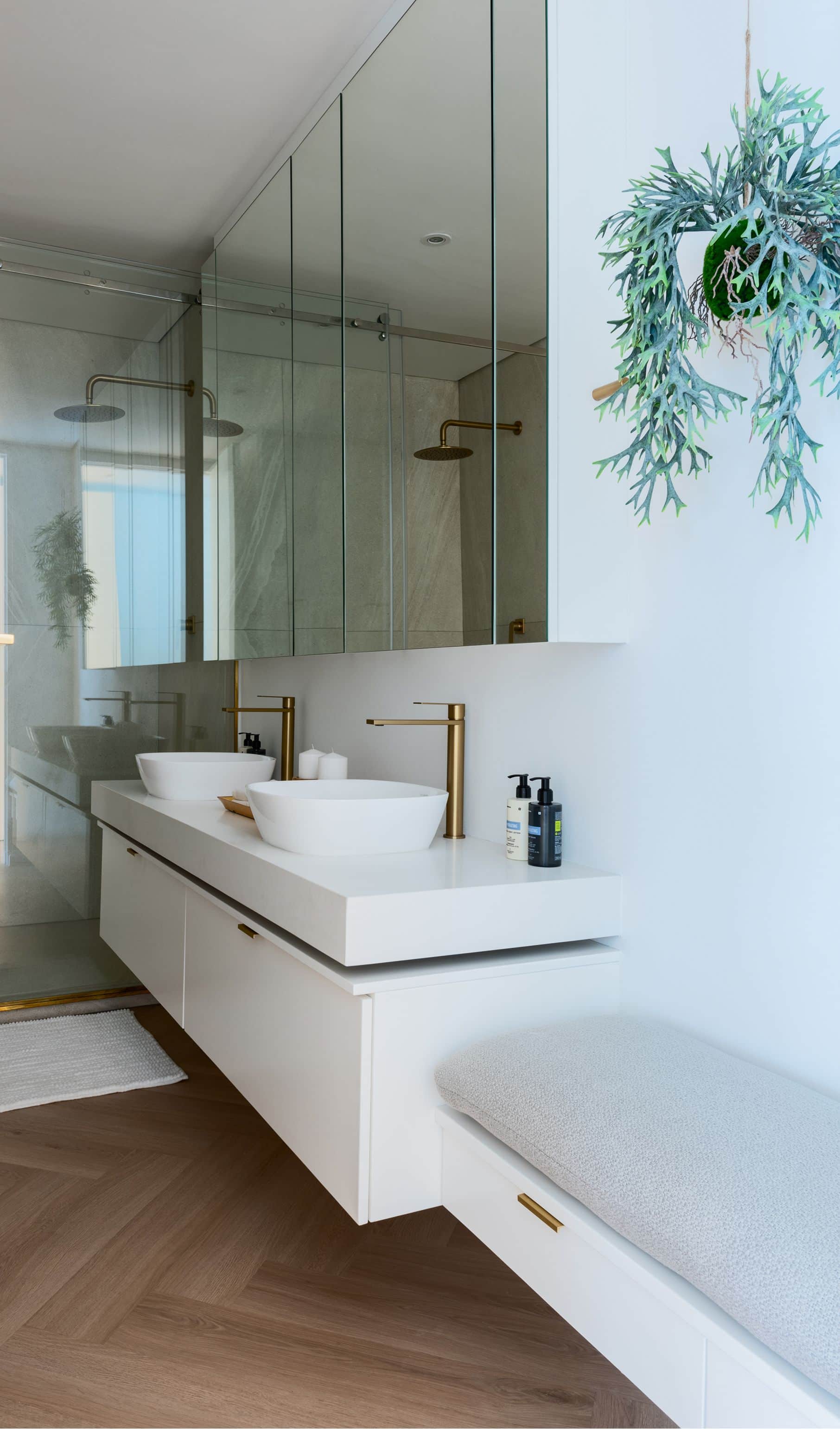 Two vessel basins with brass taps on a floating white vanity beside an upholstered bench