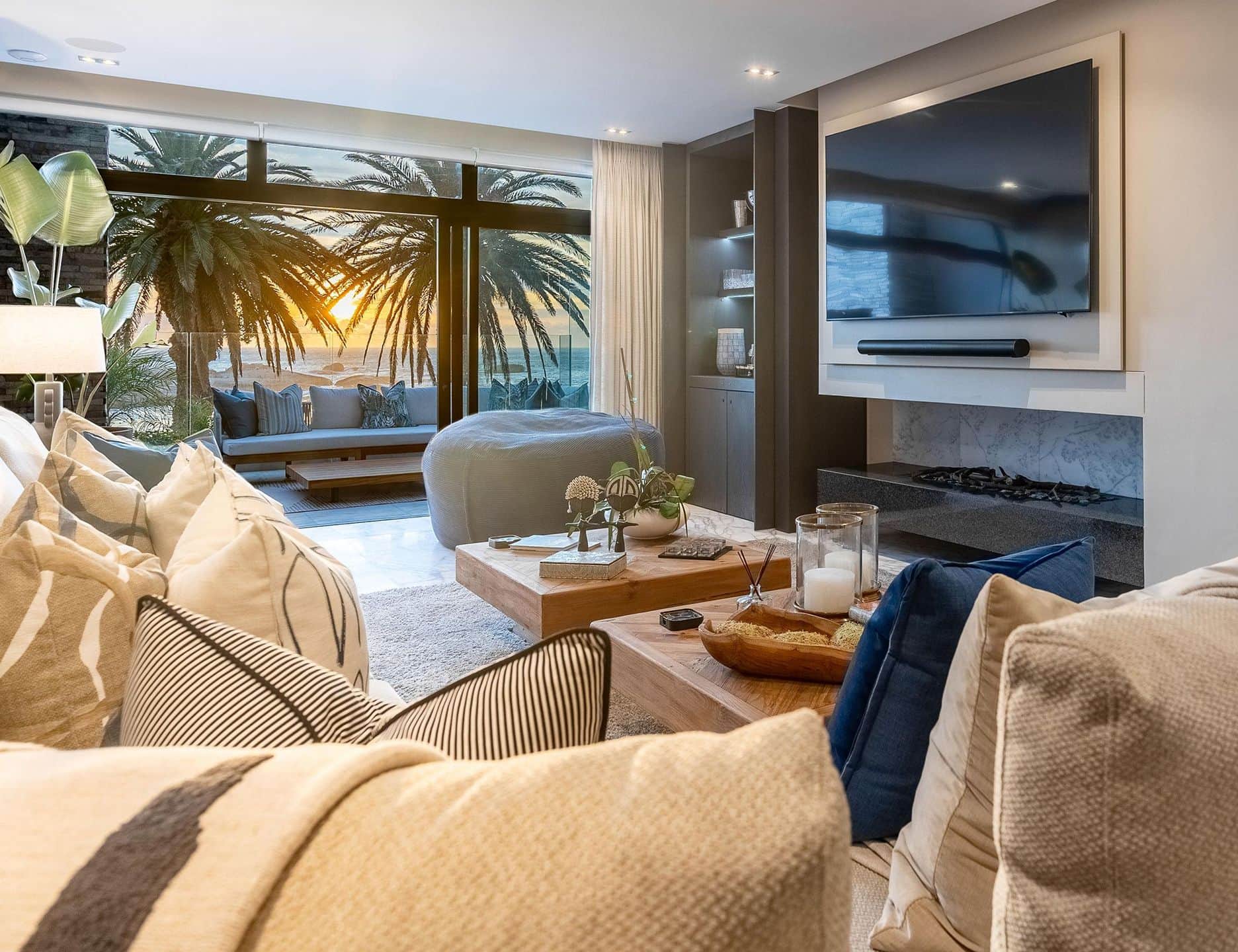 Lounge with linen sofas and a wall-mounted TV above a fireplace, palm trees at sunset beyond the glass