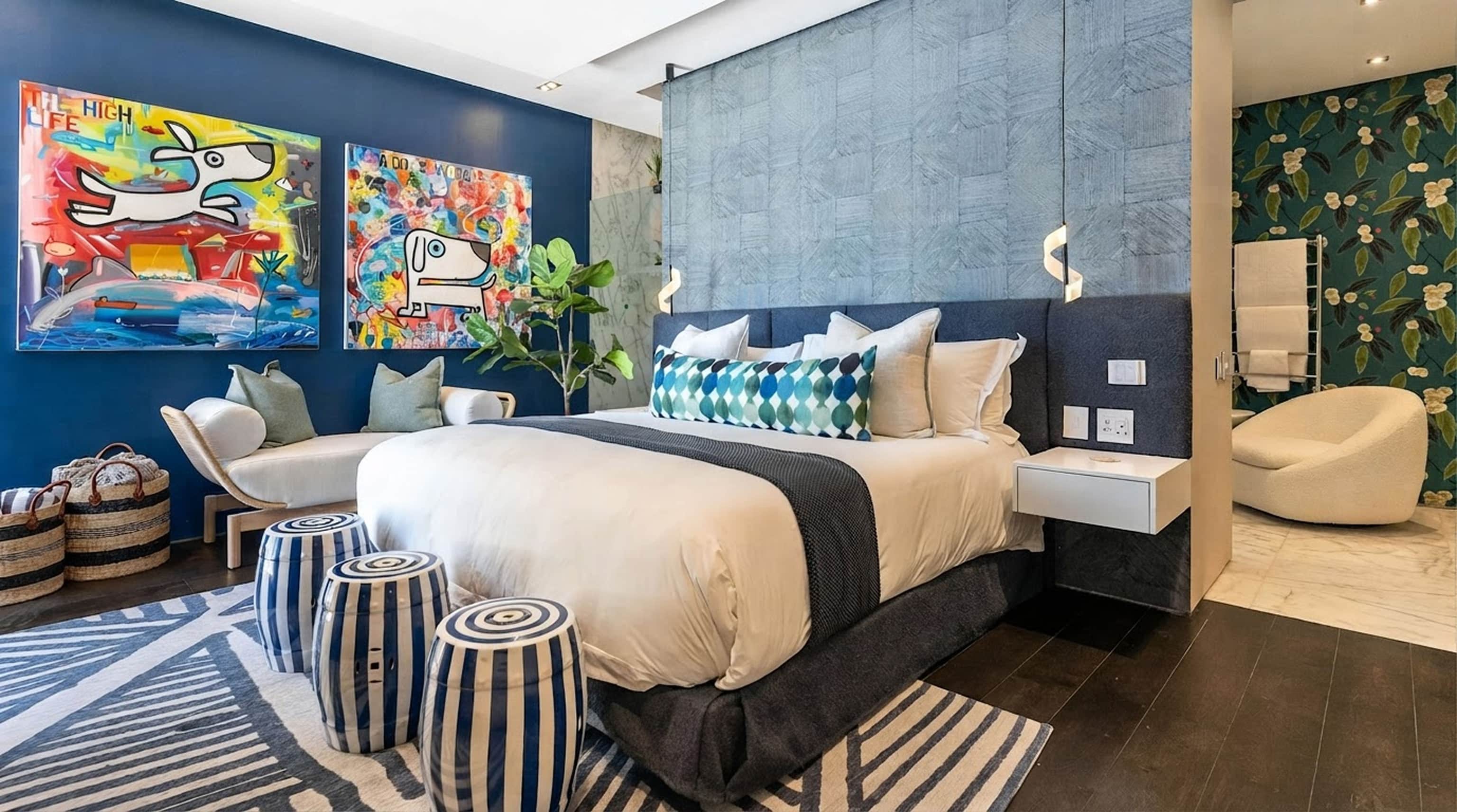 Bedroom with a navy feature wall, colourful paintings, striped stools and an en-suite with botanical wallpaper